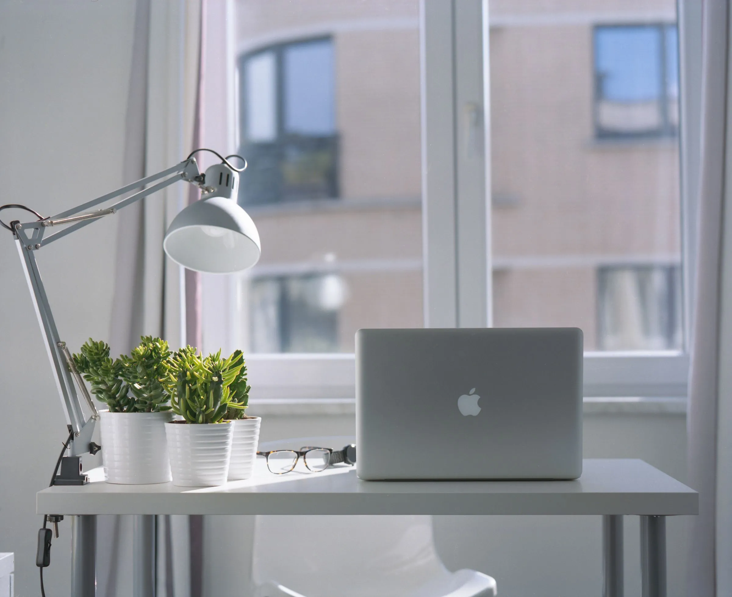 A laptop on a white desk under an angled lamp, beside potted plants.