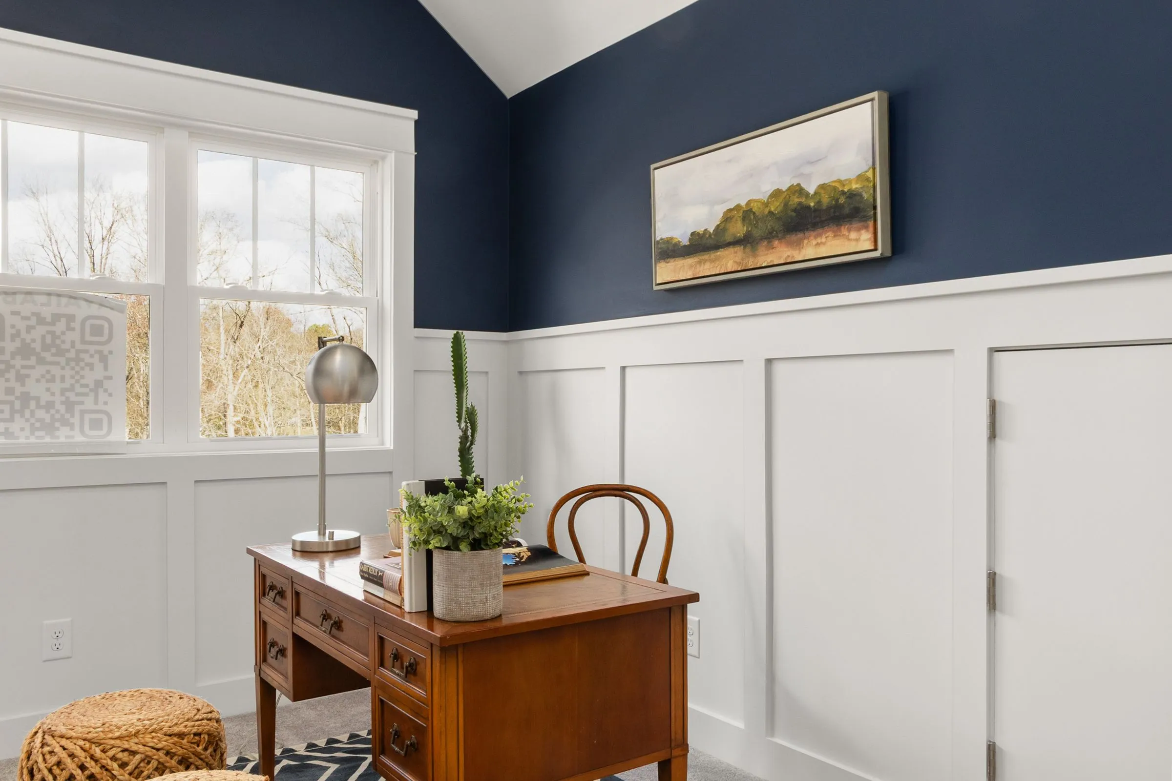 A quiet office with a wooden desk, a lamp and plants under a tall window.