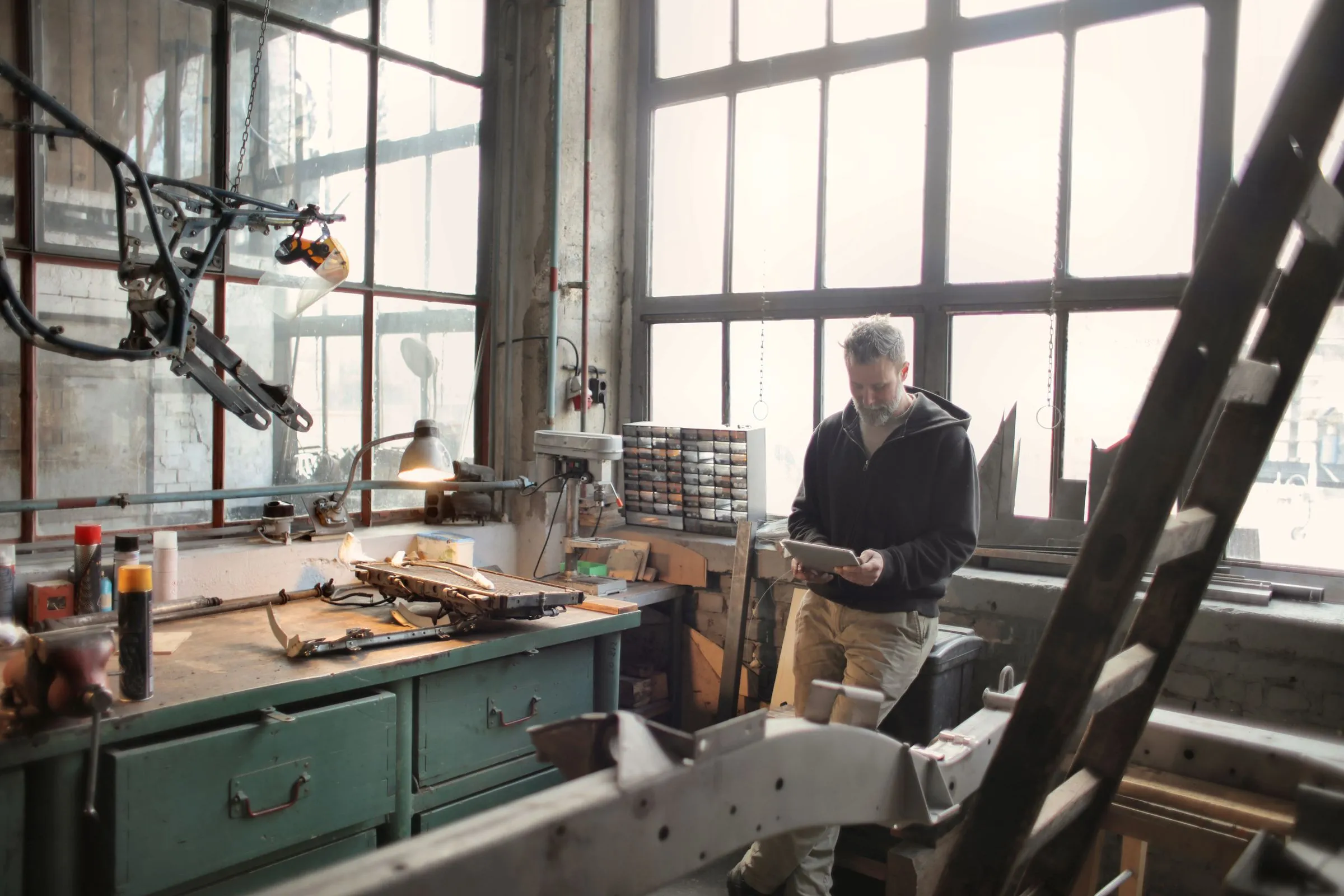 A workshop owner in a hoodie checking figures on a tablet by the tall windows of his workshop.